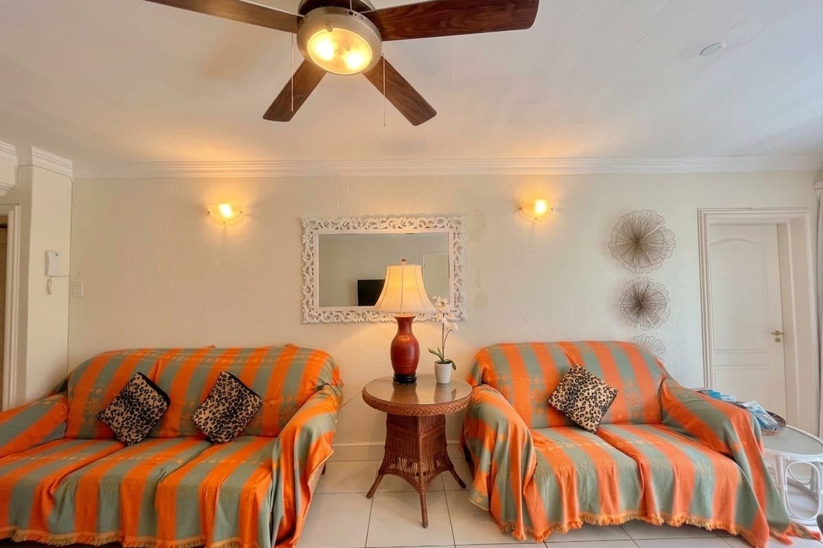 A living room featuring tiled flooring, crown molding, a ceiling fan, and wall-mounted lighting fixtures.