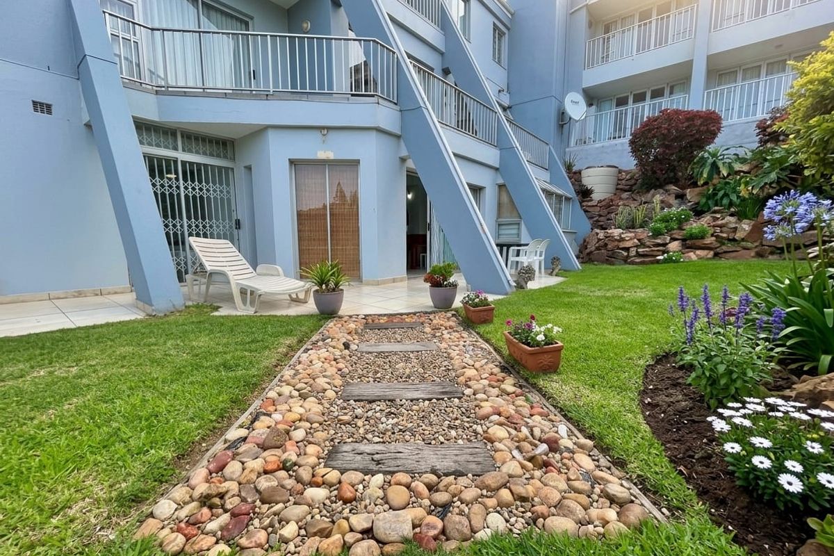 Rear view of the apartment building featuring a tiled patio, landscaped garden with a stone pathway, and upper-level balconies with metal railings.