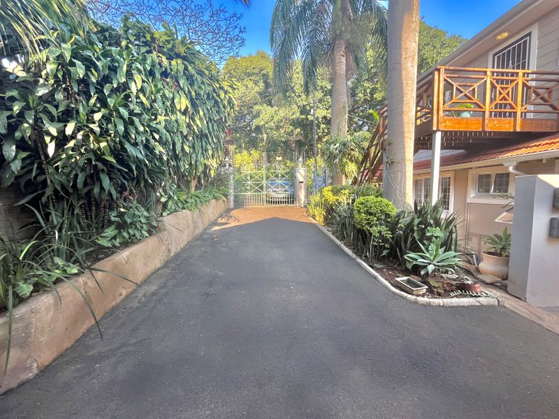A paved driveway leading to a secure metal gate, featuring a landscaped garden and an elevated wooden balcony attached to the house.