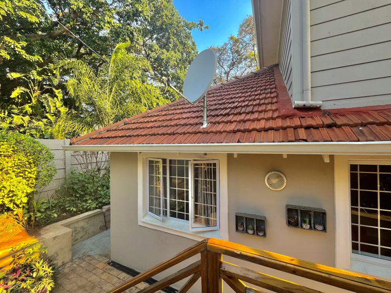 Rear exterior view of the house featuring a terracotta tiled roof, satellite dish, timber deck railing, and paved patio area with established garden surroundings.