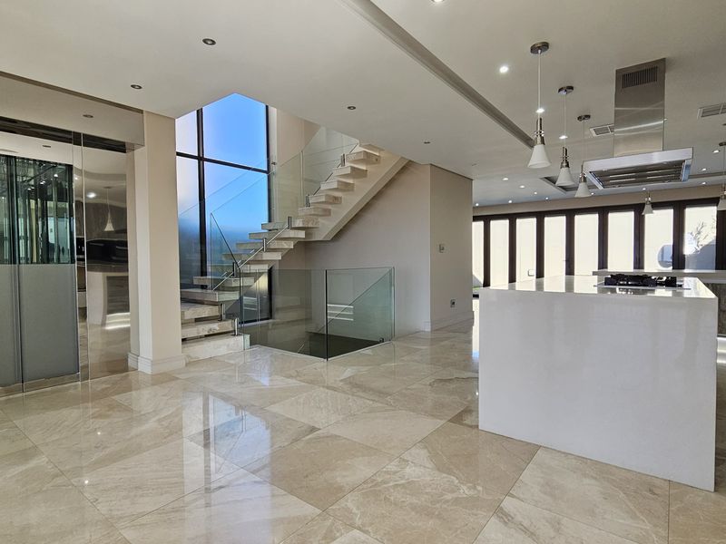 Spacious open-plan living area featuring polished stone flooring, a modern kitchen island with integrated extractor, and a striking glass-railed staircase with an internal lift.