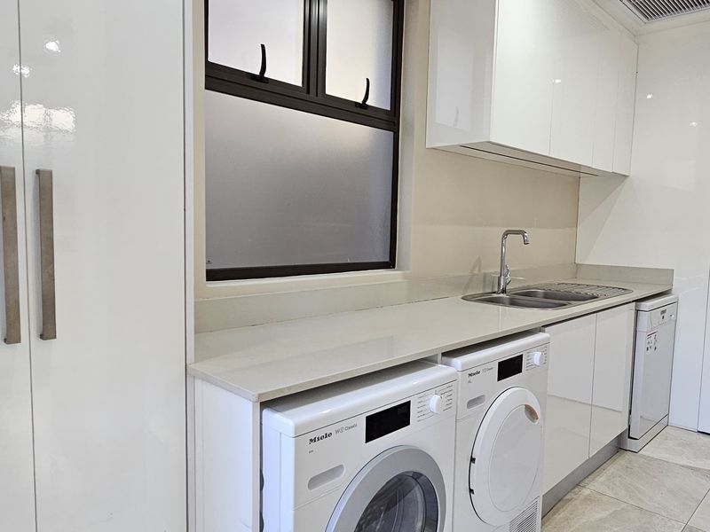 A modern, well-appointed laundry room featuring sleek white cabinetry, stone countertops, a stainless steel sink, and high-quality tiled flooring.