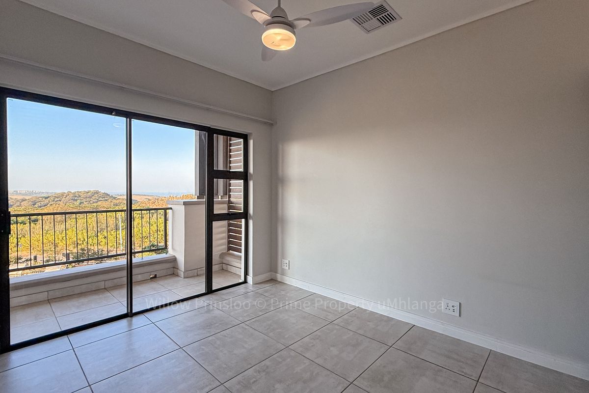 A bright, empty bedroom featuring neutral tiled floors, a ceiling fan, and large sliding glass doors that open onto a private balcony with scenic views.