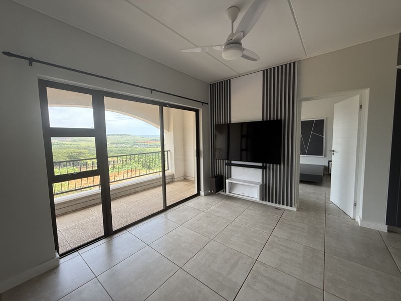 Modern living room featuring large sliding glass doors leading to a balcony, decorative wall paneling, tiled flooring, and a ceiling fan.