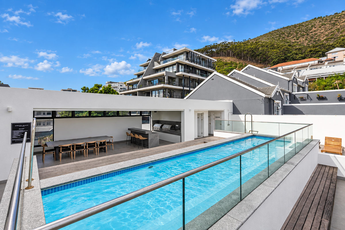 Modern rooftop pool area featuring a lap pool, glass balustrades, timber decking, and a built-in outdoor kitchen with barbecue facilities.