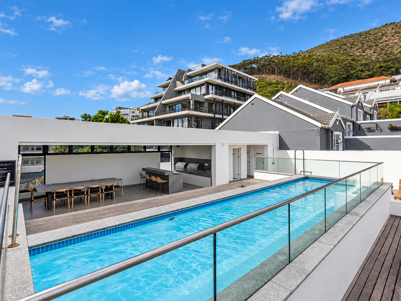 Modern rooftop pool area featuring a lap pool, glass balustrades, timber decking, and a built-in outdoor kitchen with barbecue facilities.