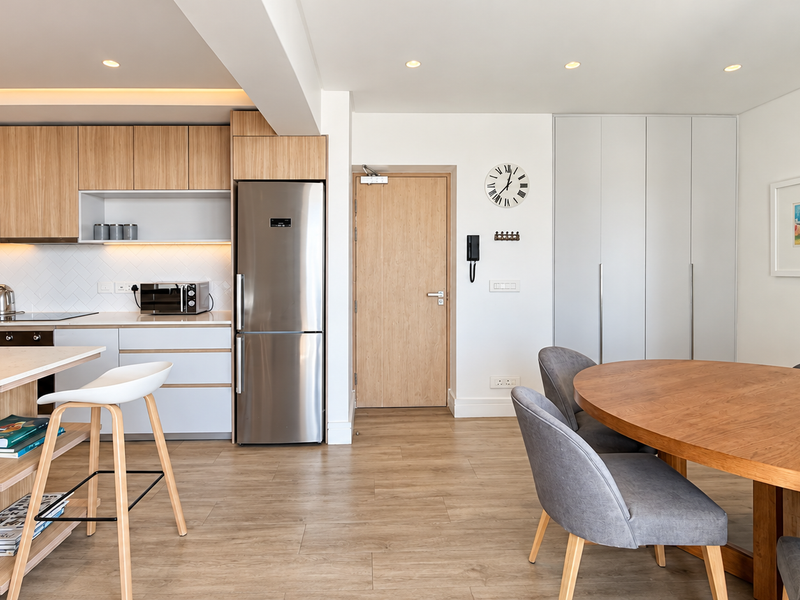 Modern open-plan kitchen and dining area featuring light wood cabinetry, stone countertops, integrated appliances, and wood-look flooring in a well-maintained apartment.