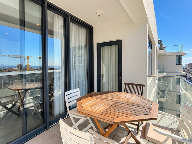 A modern, well-maintained balcony featuring a glass balustrade, tiled flooring, and large sliding glass doors offering expansive urban and coastal views.