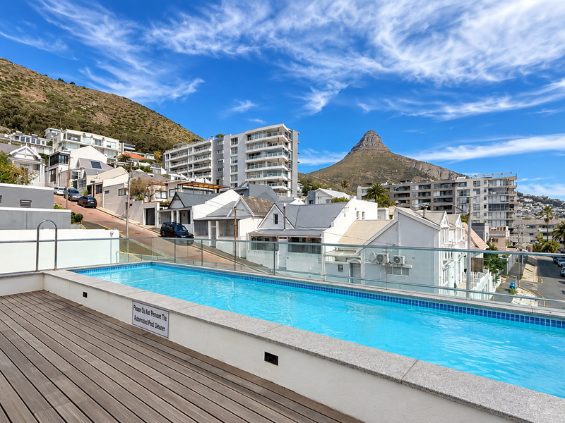 A modern elevated swimming pool featuring timber decking and a glass balustrade, offering scenic mountain and urban views.