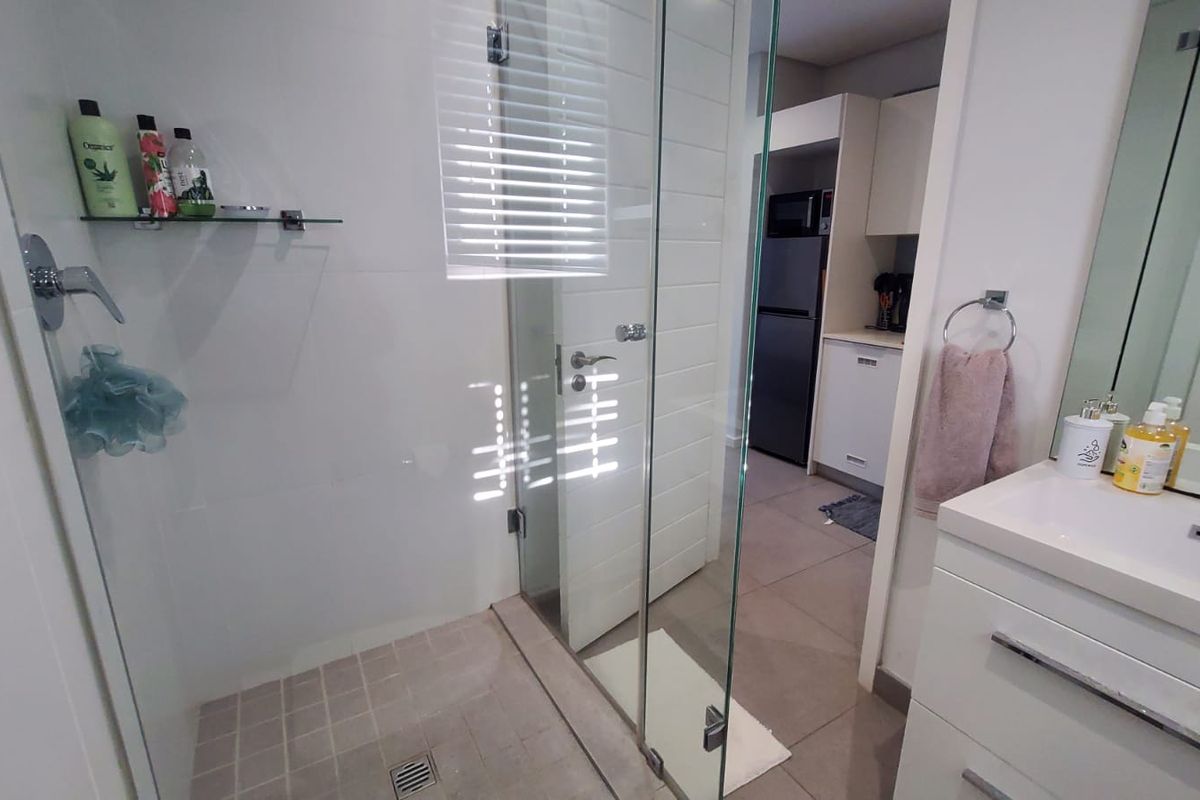 Modern bathroom featuring a glass-enclosed shower, sleek vanity with basin, and neutral tiled finishes, with a view into the adjacent kitchen area.