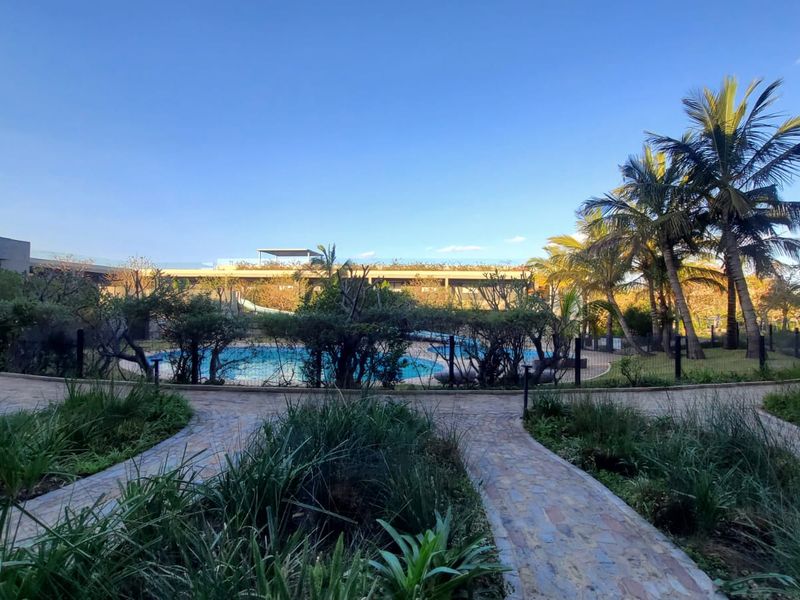 A well-maintained communal pool area featuring paved walkways, lush landscaping, and established palm trees within a secure residential complex.