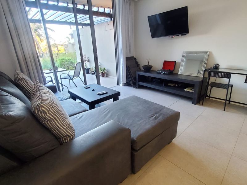 A bright living room featuring large sliding glass doors that open onto a patio, with neutral tiled flooring and a wall-mounted television.
