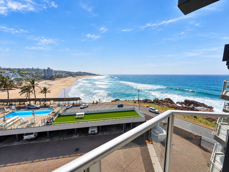 Elevated coastal view overlooking the beach, ocean, swimming pool, and paved parking area from a balcony with a glass balustrade.