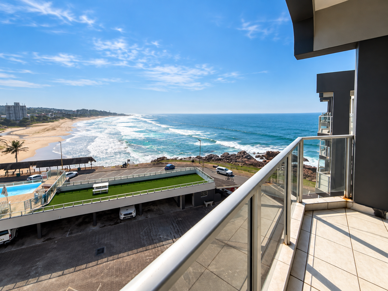 A spacious balcony featuring a modern glass balustrade and tiled flooring, offering expansive, unobstructed views of the coastline and ocean.