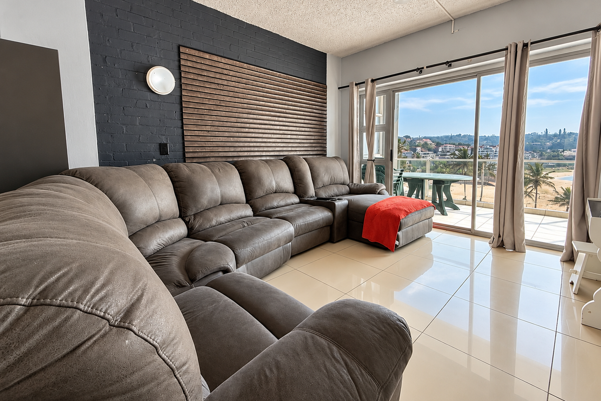 A bright living room featuring large sliding glass doors that open to a balcony with coastal views, complemented by a dark brick feature wall.