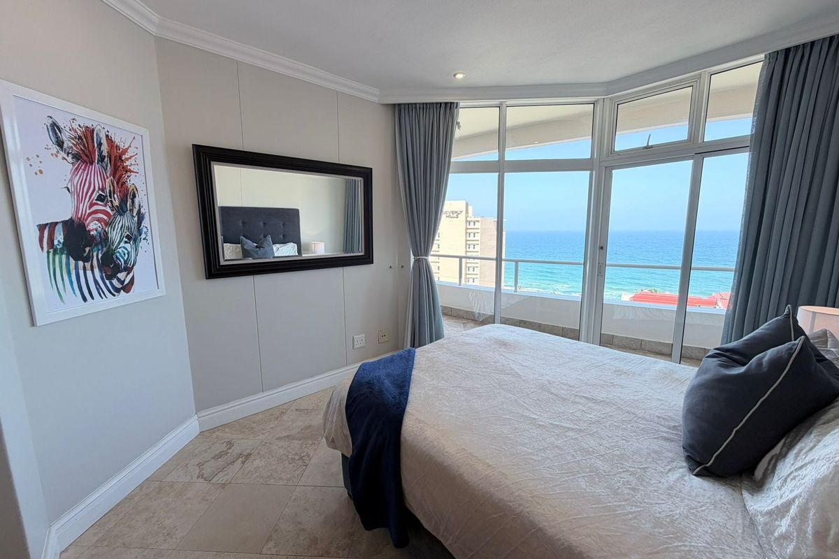 A bright bedroom featuring large floor-to-ceiling windows with direct ocean views and access to a private balcony, finished with neutral tiled flooring.