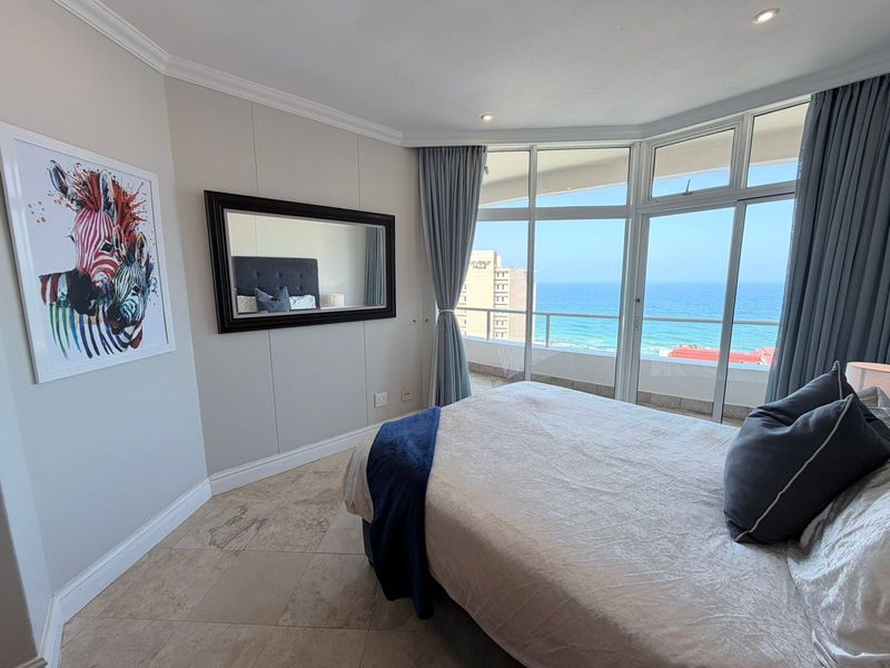 A bright bedroom featuring large floor-to-ceiling windows with direct ocean views and access to a private balcony, finished with neutral tiled flooring.