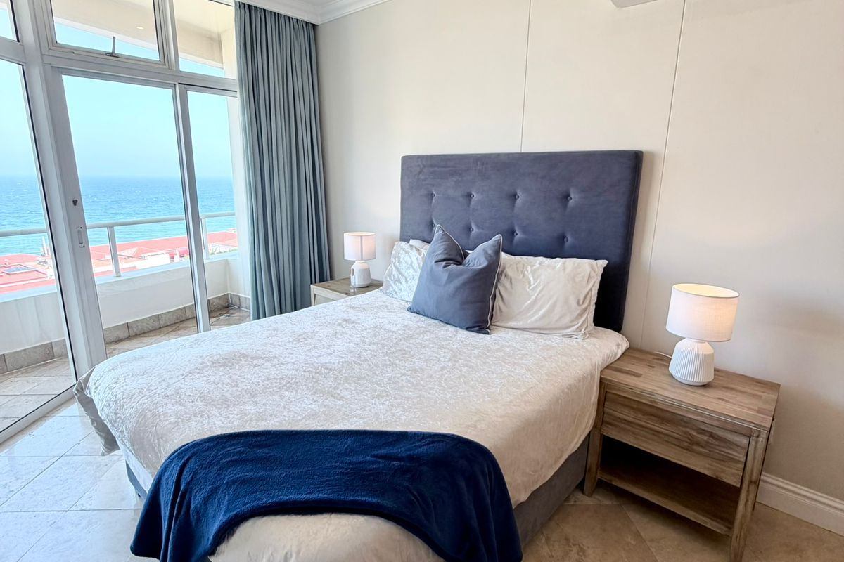 A well-maintained bedroom featuring tiled flooring, a split-system air conditioner, and large sliding glass doors providing direct access to a balcony with ocean views.