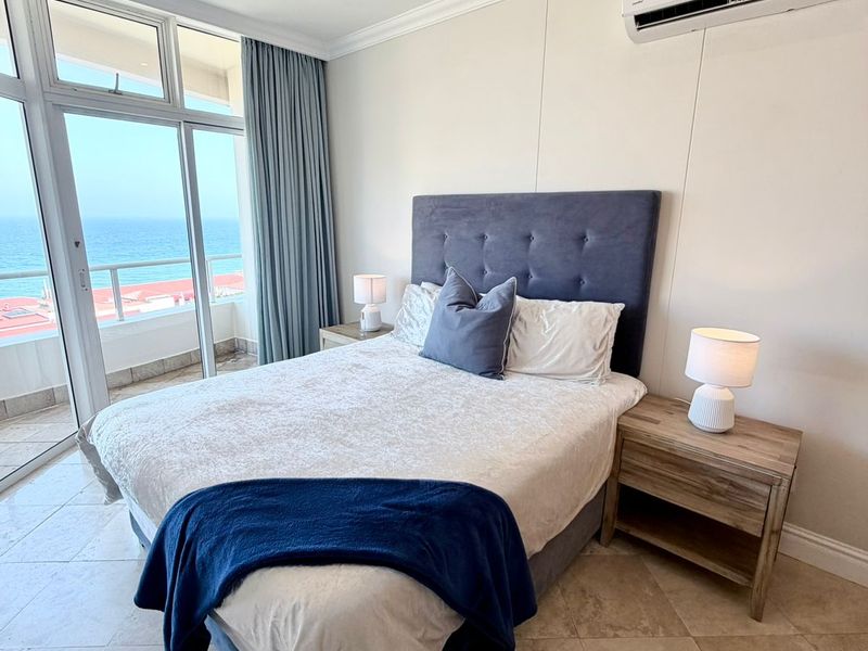 A well-maintained bedroom featuring tiled flooring, a split-system air conditioner, and large sliding glass doors providing direct access to a balcony with ocean views.