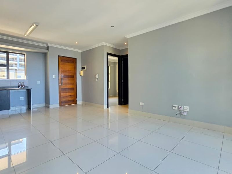 Spacious open-plan living area featuring modern tiled flooring, neutral grey walls, and a compact kitchen with built-in cabinetry.