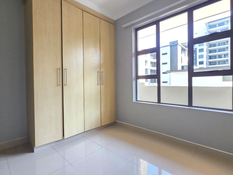 A bright, empty bedroom featuring neutral tiled flooring, light wood-finish built-in cupboards, and large aluminium-framed windows.