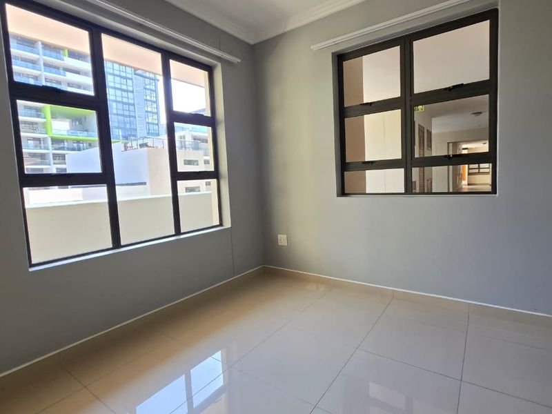 A bright, empty bedroom featuring neutral grey walls, large aluminium-framed windows, and polished floor tiles, offering a clean and modern blank canvas.