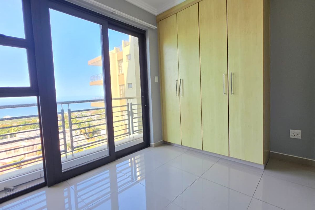 A bright, empty bedroom featuring built-in cupboards, sleek tiled flooring, and a large sliding glass door leading to a balcony with coastal views.