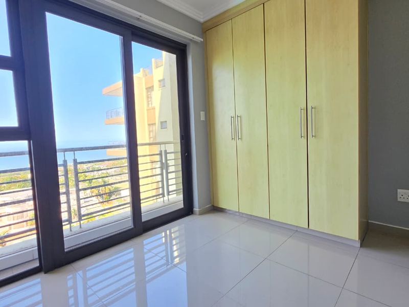 A bright, empty bedroom featuring built-in cupboards, sleek tiled flooring, and a large sliding glass door leading to a balcony with coastal views.