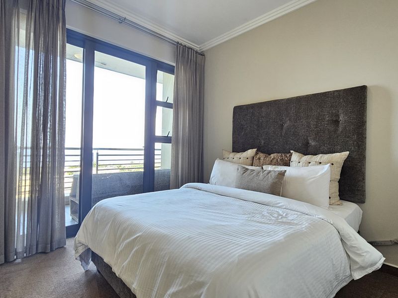 A well-lit bedroom featuring neutral tones, carpeted flooring, built-in wardrobe, and a sliding glass door providing access to a private balcony.