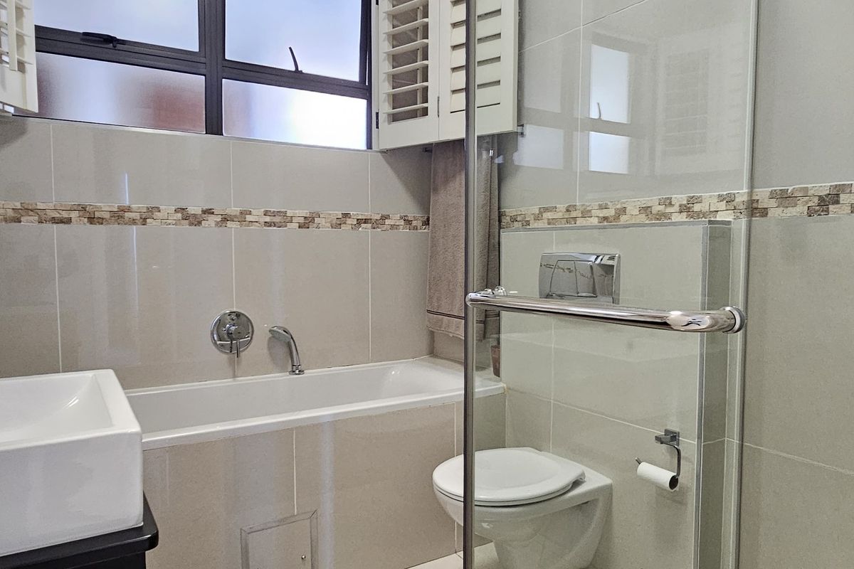 Modern bathroom featuring a glass-enclosed shower, built-in bathtub, wall-mounted toilet, and neutral tiled finishes with shutter window detailing.