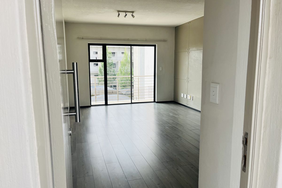 An empty, well-lit living area featuring modern laminate flooring, built-in wall paneling, and a sliding glass door leading to a private balcony.