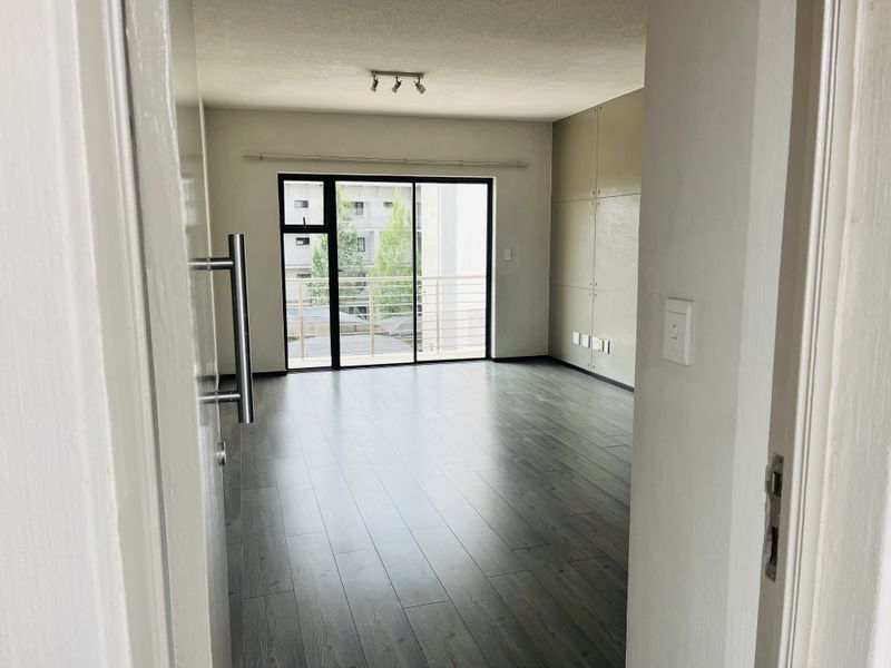 An empty, well-lit living area featuring modern laminate flooring, built-in wall paneling, and a sliding glass door leading to a private balcony.