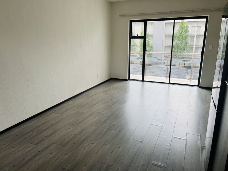 A bright, empty living room featuring modern laminate flooring and a large sliding glass door providing access to a private balcony.