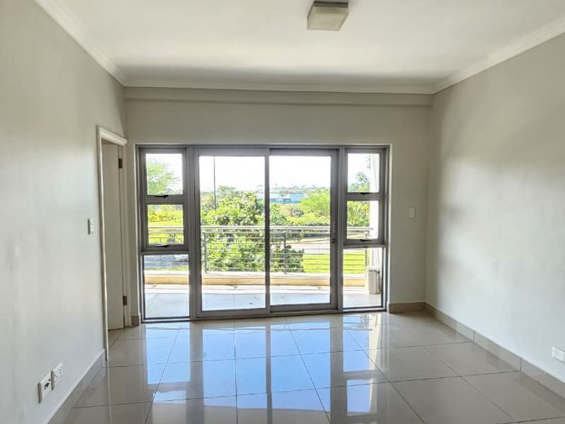 A bright, empty living room featuring neutral tiled flooring and a large sliding glass door that opens onto a balcony with outdoor views.