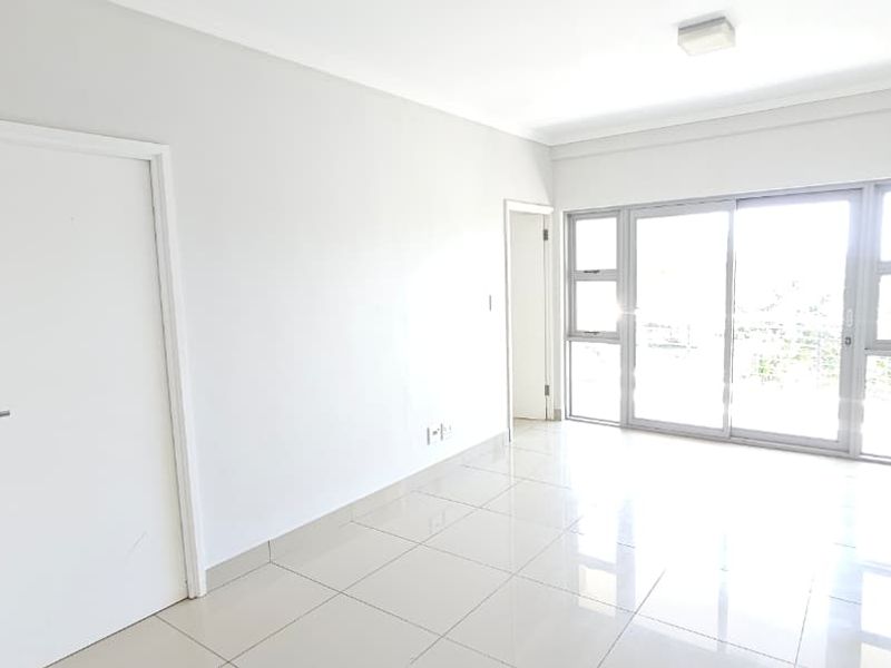 A bright, empty living area featuring neutral tiled flooring, white walls, and a large sliding glass door providing natural light and access to the exterior.