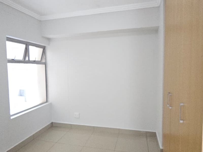A bright, empty bedroom featuring neutral tiled flooring, built-in wooden cupboards, and a large window providing natural light.