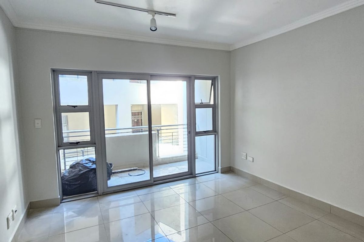 An empty living room featuring neutral tiled flooring, cove cornicing, and a large sliding glass door providing access to a balcony.