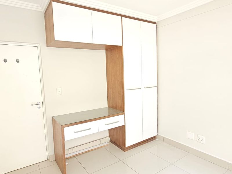 A clean, empty bedroom featuring a modern built-in wardrobe and integrated desk unit with white and wood-grain finishes and tiled flooring.