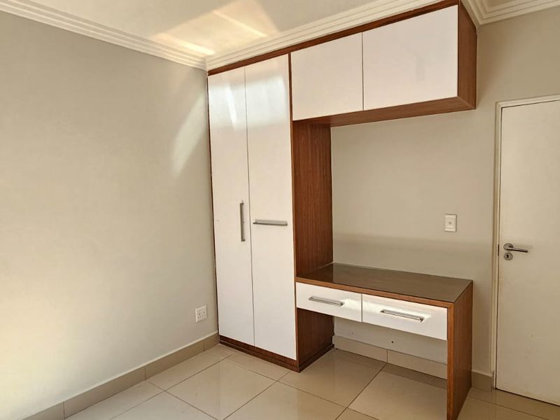 A clean, empty bedroom featuring modern built-in cabinetry with a desk, overhead storage, and neutral tiled flooring.