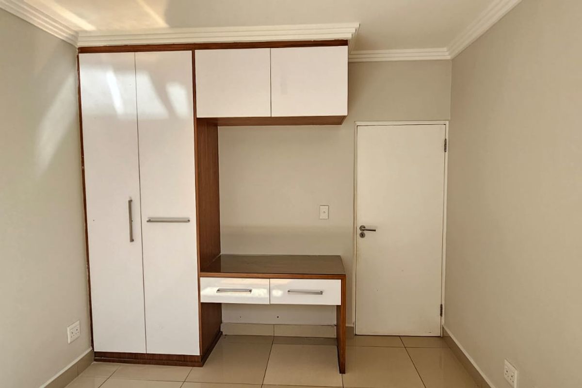 A clean bedroom featuring a modern built-in wardrobe with integrated desk space, overhead storage, and neutral tiled flooring.