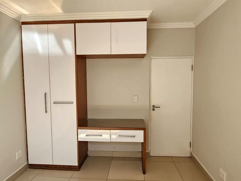 A clean bedroom featuring a modern built-in wardrobe with integrated desk space, overhead storage, and neutral tiled flooring.