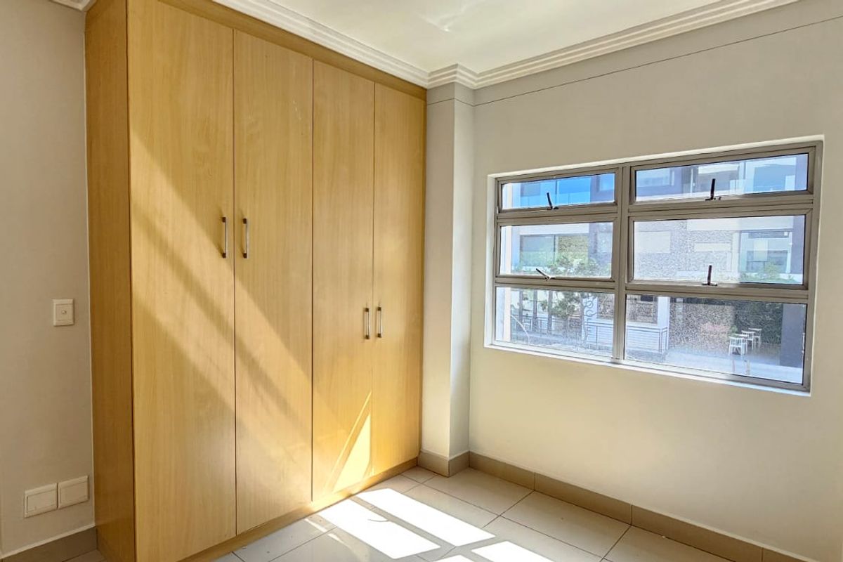 A bright, empty bedroom featuring light wood-finish built-in cupboards, tiled flooring, and large aluminium-framed windows providing natural light.