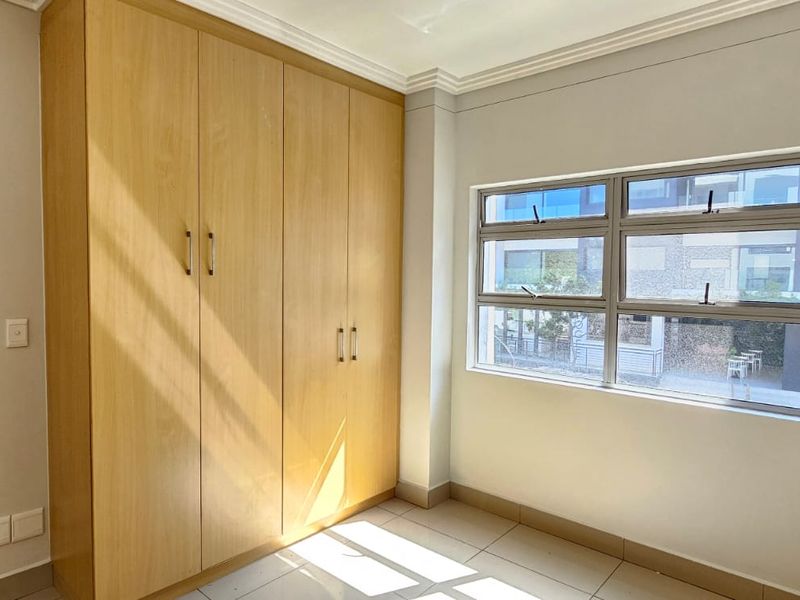 A bright, empty bedroom featuring light wood-finish built-in cupboards, tiled flooring, and large aluminium-framed windows providing natural light.