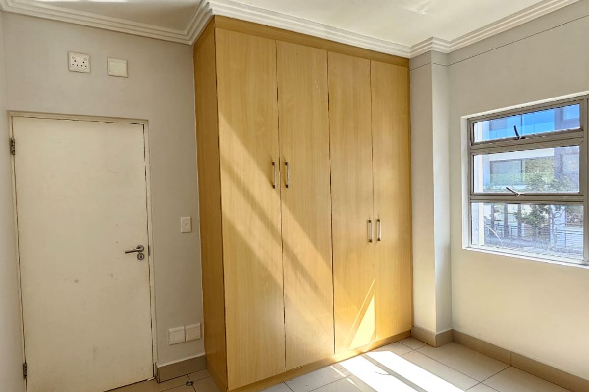 A bright, empty bedroom featuring built-in wooden cupboards, tiled flooring, and large aluminium-framed windows providing natural light.