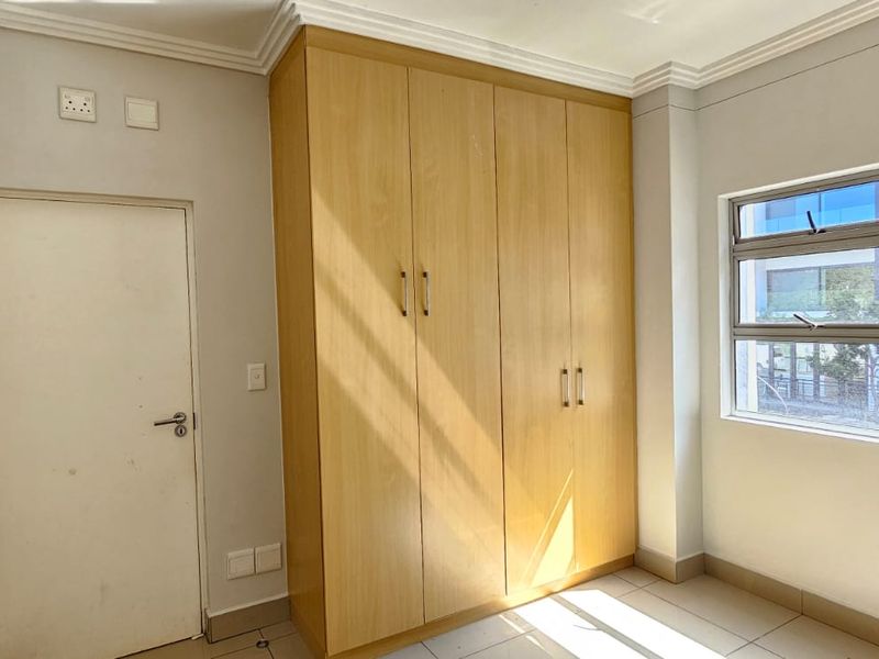 A bright, empty bedroom featuring built-in wooden cupboards, tiled flooring, and large aluminium-framed windows providing natural light.