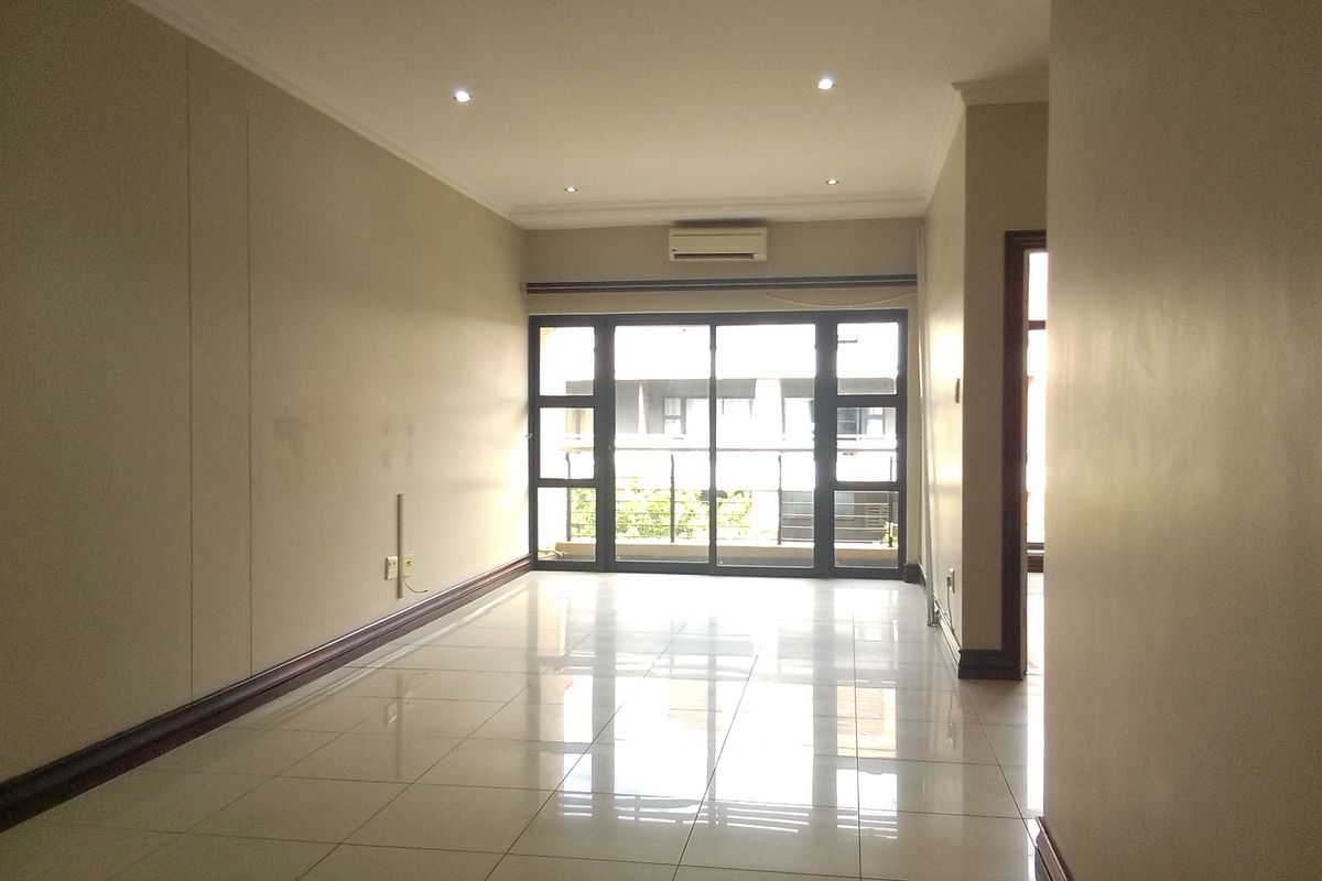 A spacious, empty living area featuring neutral tiled floors, recessed lighting, and a wall-mounted air conditioning unit with access to a balcony.