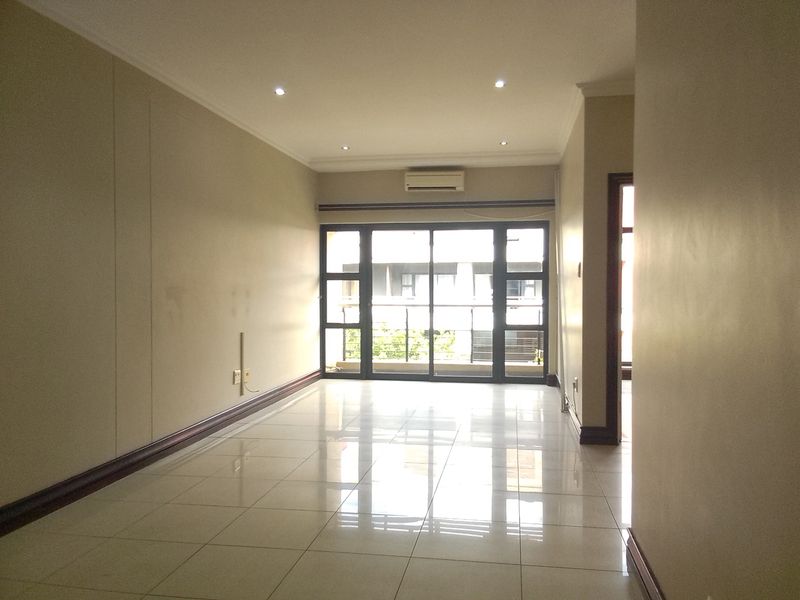 A spacious, empty living area featuring neutral tiled floors, recessed lighting, and a wall-mounted air conditioning unit with access to a balcony.