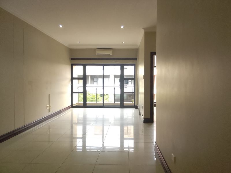 A bright, empty living room featuring polished ceramic floor tiles, recessed lighting, a wall-mounted air conditioning unit, and large sliding doors leading to a balcony.