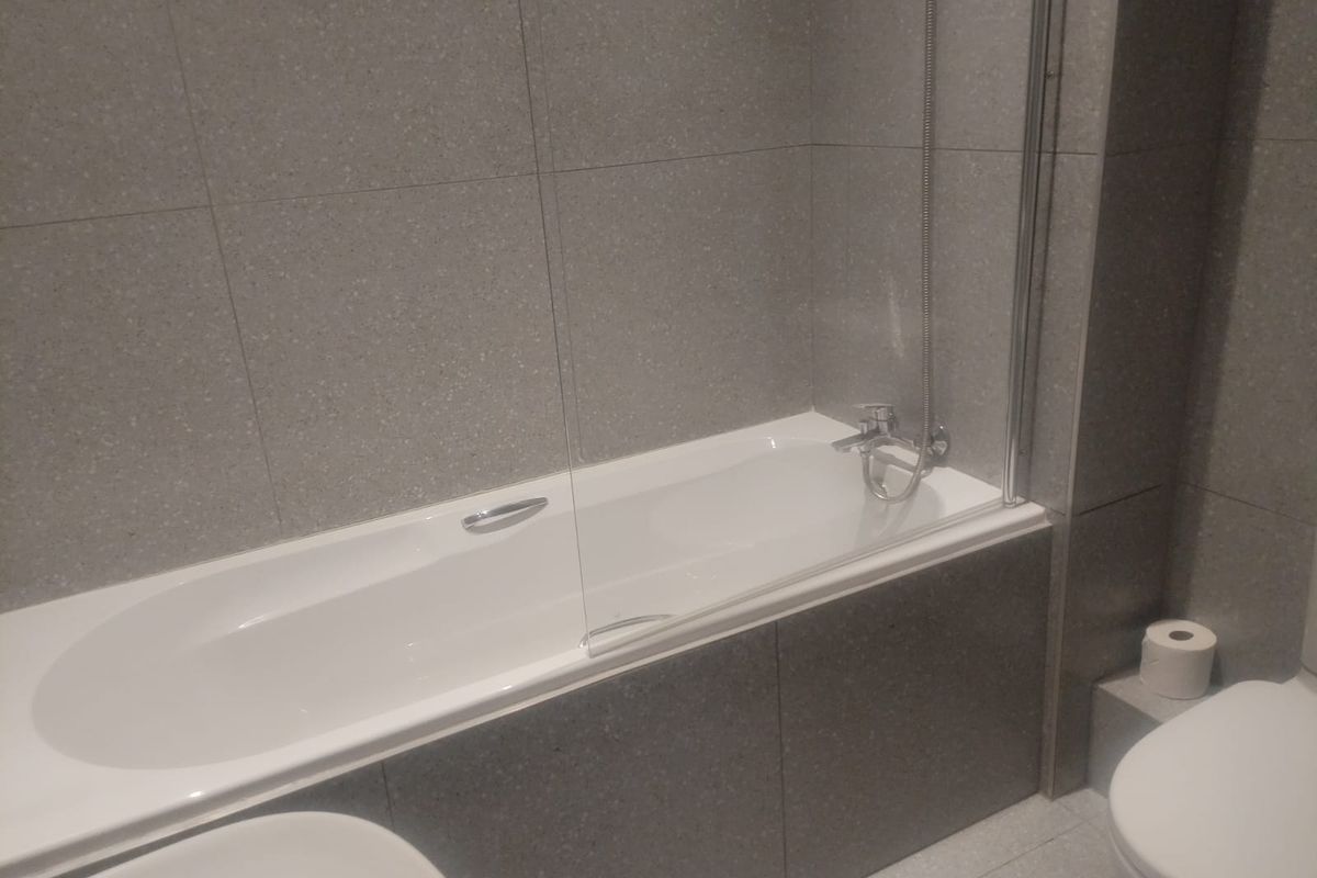 A modern bathroom featuring grey tiled walls and flooring, a built-in bathtub with a glass shower screen, and a standard toilet.