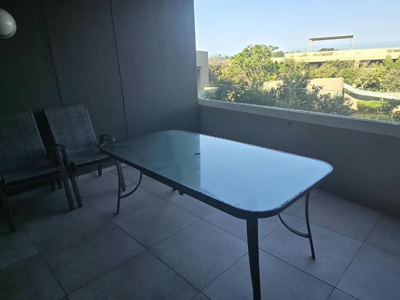 A covered balcony featuring tiled flooring, a glass balustrade, and a wall-mounted light fixture, offering an outdoor space for relaxation.